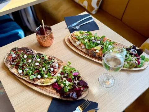 Deux pinsas garnies de roquette servies sur planche, avec un cocktail en tasse cuivrée